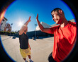 © Studio Marmellata - Two young men, one in a red shirt and one in black, exchanging a high-five outdoors with a fisheye lens capturing the vibrant sunny day and urban setting in the background.