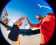 © Studio Marmellata - Two young men, one in a red shirt and one in black, exchanging a high-five outdoors with a fisheye lens capturing the vibrant sunny day and urban setting in the background.