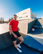 © Studio Marmellata - Male runner in a bright red shirt leans casually against a modern architectural structure under a clear blue sky on a sunny day, exuding confidence and style in a contemporary setting.