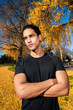 © Studio Marmellata - man in a black shirt posing confidently with arms crossed amid a vibrant autumn scene featuring golden leaves, tall trees, and a bright blue sky creating a striking contrast.