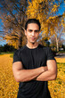 © Studio Marmellata - man in a black shirt posing confidently with arms crossed amid a vibrant autumn scene featuring golden leaves, tall trees, and a bright blue sky creating a striking contrast.