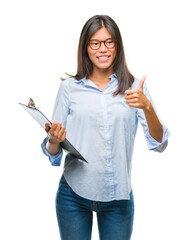 Wall Mural - Young asian business woman over isolated background holding clipboard happy with big smile doing ok sign, thumb up with fingers, excellent sign