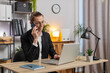 © Andrii Iemelianenko - Happy Caucasian stylish young businessman communicating with customer by phone smartphone, working on laptop netbook at home office desk. Guy in suit represents concept of distance job on Internet.
