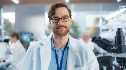 Leinwandbilder - Portrait of a Bearded Young Lab Engineer in White Coat Looking at Camera and Smiling. Medical Research Scientist Looking at Biological Samples Under a Microscope in an Applied Science Laboratory