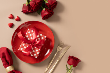 Valentine's day table setting with red plate, chocolate sweets, romantic gift and red roses on beige background. View from above.