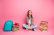 © deagreez - Adorable young girl with blonde hair surrounded by books and backpack, sitting on a pink background, looking cheerful and trendy