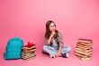 © deagreez - Thoughtful young girl sitting amidst books and a backpack on a pink background, reflecting on learning and school concepts.