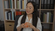 © Krakenimages.com - Asian woman wearing glasses and headset holds stress ball while sitting in an office with bookshelves in the background, illustrating a calm work environment.