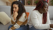 © Krakenimages.com - Women sitting on couch appearing thoughtful and distant in cozy living room environment showcasing complex friendship dynamics indoors.