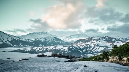 Fototapete - stunning mountainous landscape covered in snow, with a mix of green and white colors. The sky above is partly cloudy, adding depth and contrast to the scene.