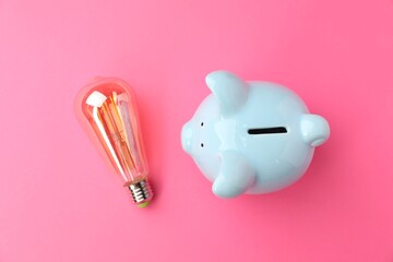 Wall Mural - Piggy bank and light bulb on pink background, flat lay. Energy saving concept