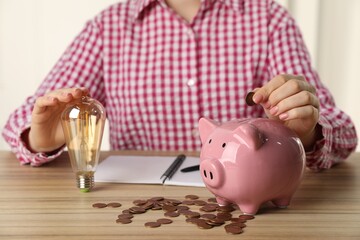 Wall Mural - Woman with light bulb putting coin into piggy bank at wooden table, closeup. Energy saving concept