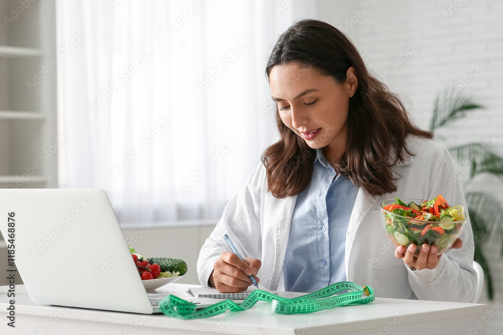 Young female nutritionist with vegetable salad writing on notebook at table in office