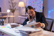 © crizzystudio - Asian businesswoman working late in office using laptop and talking on phone while reviewing charts and graphs in documents, focused on work and problem-solving