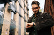 © dikushin - Low-angle portrait of fashionable young man in brown leather jacket checking smartwatch outside modern building, possibly during workday commute or break. Concept of modern successful city lifestyle.