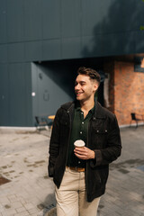 Wall Mural - Vertical portrait of cheerful young man in casual clothes enjoying coffee break, holding takeaway cup while walking down city street on sunny day, radiating sense of urban leisure and relaxation.