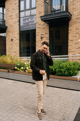 Wall Mural - Full length vertical portrait of stylish man in casual clothes holding paper cup of coffee and talking on smartphone while walking down street by modern brick building. Concept of urban lifestyle.