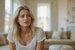 © polack - Unhappy woman with blonde hair and blue eyes is sitting on a couch in a living room, she looks sad and is staring off into the distance