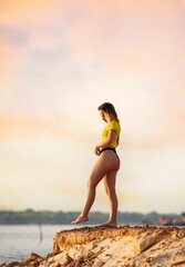 Wall Mural - young brazilian woman in a yellow shirt standing on sandy cliff at sunset