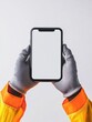© Ivan - A pair of hands wearing gray protective gloves holds a smartphone with an empty screen. The individual is wearing a bright orange safety jacket