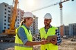 © miss irine - Two cheerful construction workers wearing hard hats, safety vests discuss project details on tablet outdoors at building site. Workers happy, focused on building project. Modern tech tools used at