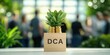 © Pixelbiz - DCA wooden block concept. A small plant in a light-colored pot labeled 'DCA' is prominently featured, with a blurred background of people and greenery.