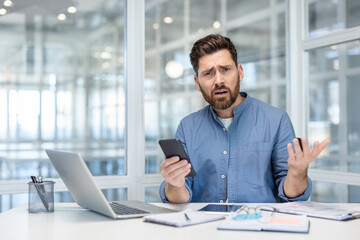 upset and disappointed man with phone in hands. office worker at workplace looking anxiously at came