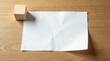 © Sumrit - A simple wooden cube rests beside a subtly creased sheet of blank paper on a light brown wooden surface.