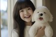 © Bay - A young girl with long hair smiles brightly as she holds a fluffy white dog, conveying a sense of joy and affection in a warm indoor setting