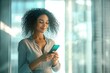 © Yuliya - Smiling business woman holding smartphone in modern office space with natural light