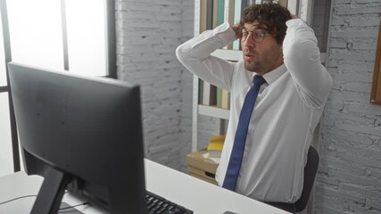 Wall Mural - Young man in office wearing glasses and tie, appears stressed with hands on head, sitting at desk with computer, bookshelf in background, indicating frustration at work.