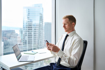 Wall Mural - Busy professional business man sitting in office holding smartphone. Young businessman executive using ai apps on cellphone working on laptop computer technology looking at mobile phone at work.