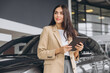 © anatoliycherkas - Smiling car seller, businesswoman in beige suit standing in car salon and using tablet for choosing right car.