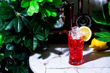 Sticker - Bitter lemon cooler alcoholic cocktail drink with gin, dry vermouth, juice, grenadine, soda, ice and lemon. Marble table in lounge bar with green plants