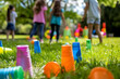 © Rich4289 - Colorful plastic cups scattered on grass with children playing in background, enjoying sunny day outdoors