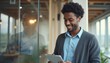 © natakot - A cheerful Black man in a cozy cardigan smiles while using a tablet in a stylish office setting. The warm ambiance and reflections in the glass create a vibrant atmosphere of productivity and joy.