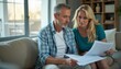 © natakot - A middle-aged couple, a man with gray hair and a woman with long blonde hair, sit closely on a cozy sofa, intently reviewing important documents. Their focused expressions convey a sense of