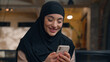 © Yuliia - Muslim woman in hijab islamic girl Arabian businesswoman entrepreneur calm smiling business lady working at desk scrolling mobile phone gadget addicted female online app cellphone internet in office
