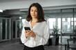 © Stock 4 You - Young Indian business woman manager ceo using cell phone mobile app laptop. Smiling Latin Hispanic mature female businesswoman holding smartphone in office trading, reading, working online. Copy space