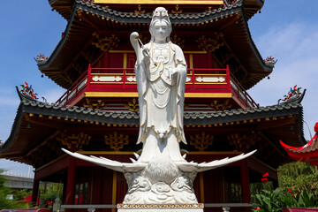 A detailed close-up of a magnificent white Guan Yin statue, positioned in front of a vibrant red and gold Chinese pagoda.