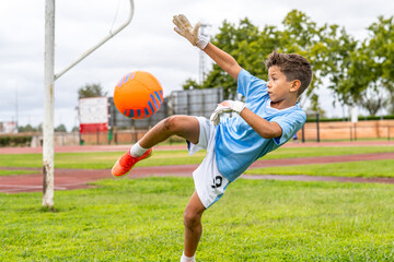 Wall Mural - Young soccer goalkeeper training, wearing protective gloves while powerfully kicking orange and blue ball during intense practice session