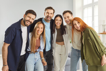 Group of happy smiling young people friends or colleagues in casual clothes looking cheerful and positively at camera indoors. Portrait of joyful men and women standing in a row together.