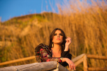 Wall Mural - Spanish woman, brunette and beautiful, dancing flamenco with typical flamenco dancing costume leaning on a wooden railing and looking at the sea at the horizon at sunset. The woman is sad.