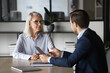 © fizkes - Two colleagues discussing project marketing strategy in office. Professional insurance agent holds paper explains policy terms and conditions with female client seated at table. Negotiations, meeting