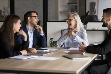 Canvas Print - Laughing businesspeople take part in briefing event in boardroom. Mature 55s and millennial 35s office employees lead talk seated at conference table, looks happy, giggling while negotiate at meeting