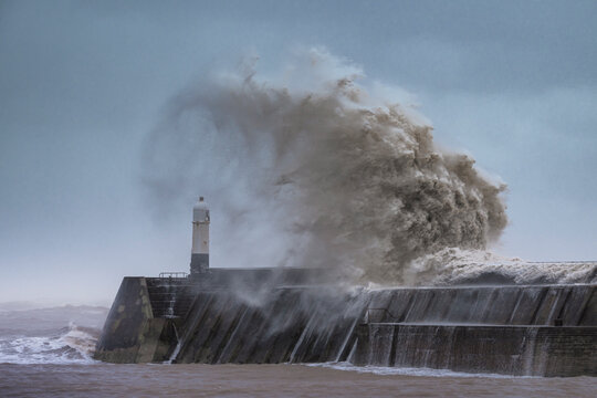 A huge winter storm hitting the coast of the UK. Giant waves crash into a breakwater, with a lighthouse. The sky is grey and moody