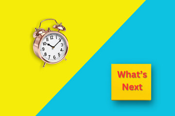 A rose gold alarm clock and a yellow sticky note with 'What’s Next' on a blue and yellow split background, representing time and future goals.