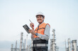 © PhugunStock - Smiling Asian male engineer wearing safety uniform standing in front of oil refinery with tablet device Radio communication to monitor oil refinery operations Petrochemical Gas Industry Engineer