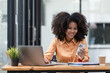 © Songsak C - Portrait of a beautiful confident businesswoman using a laptop computer holding a mobile phone sitting in a modern office. Smiling African American freelancer working online from home.
