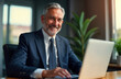 © Maryna - Smiling mature businessman works on laptop in office. Business executive uses computer for work. Pro looks happy, focused. Man in business suit sits at desk. Modern office setting. Business concept.
